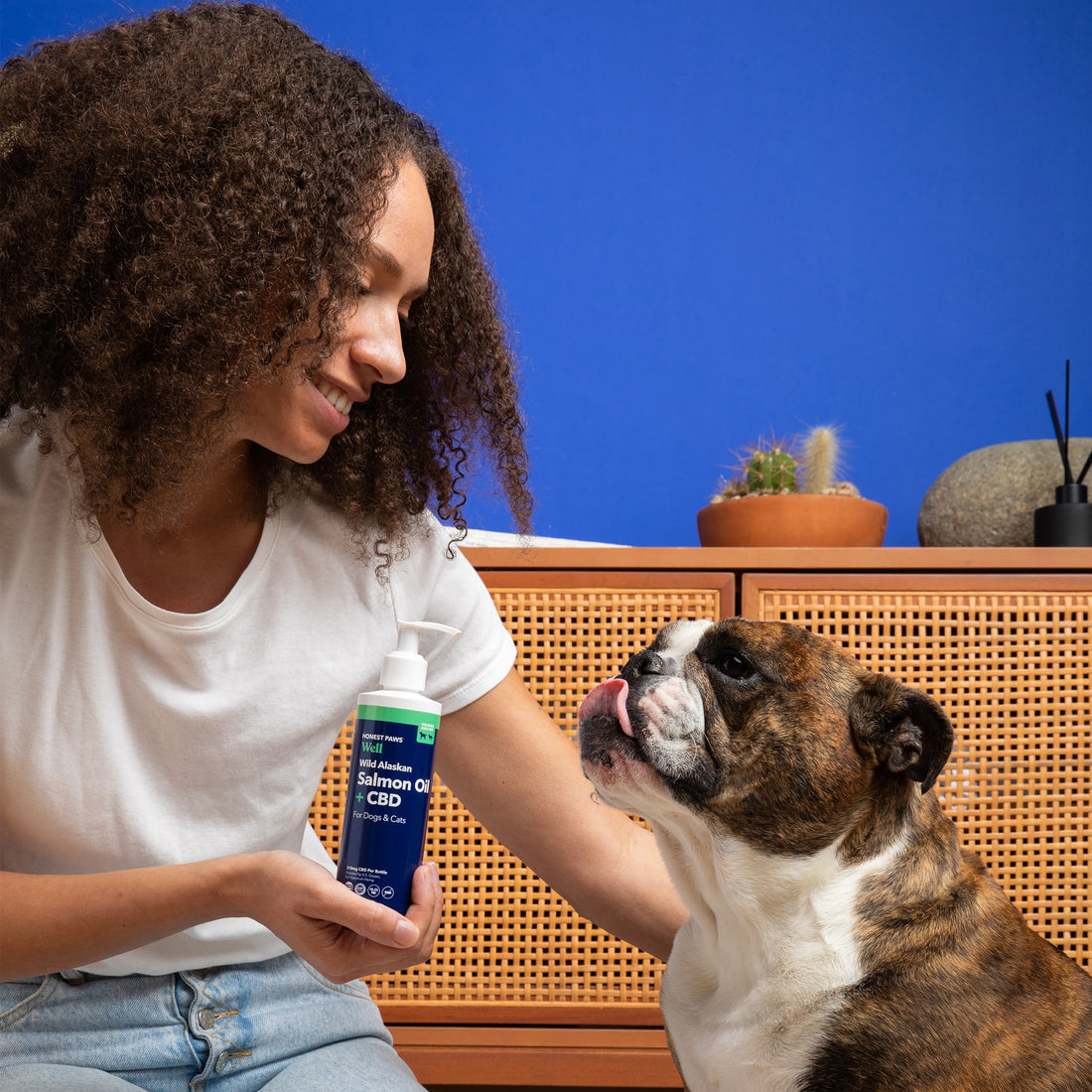 woman with brown curly hair holding bottle of salmon oil with cbd while bulldog looks at her licking his lips